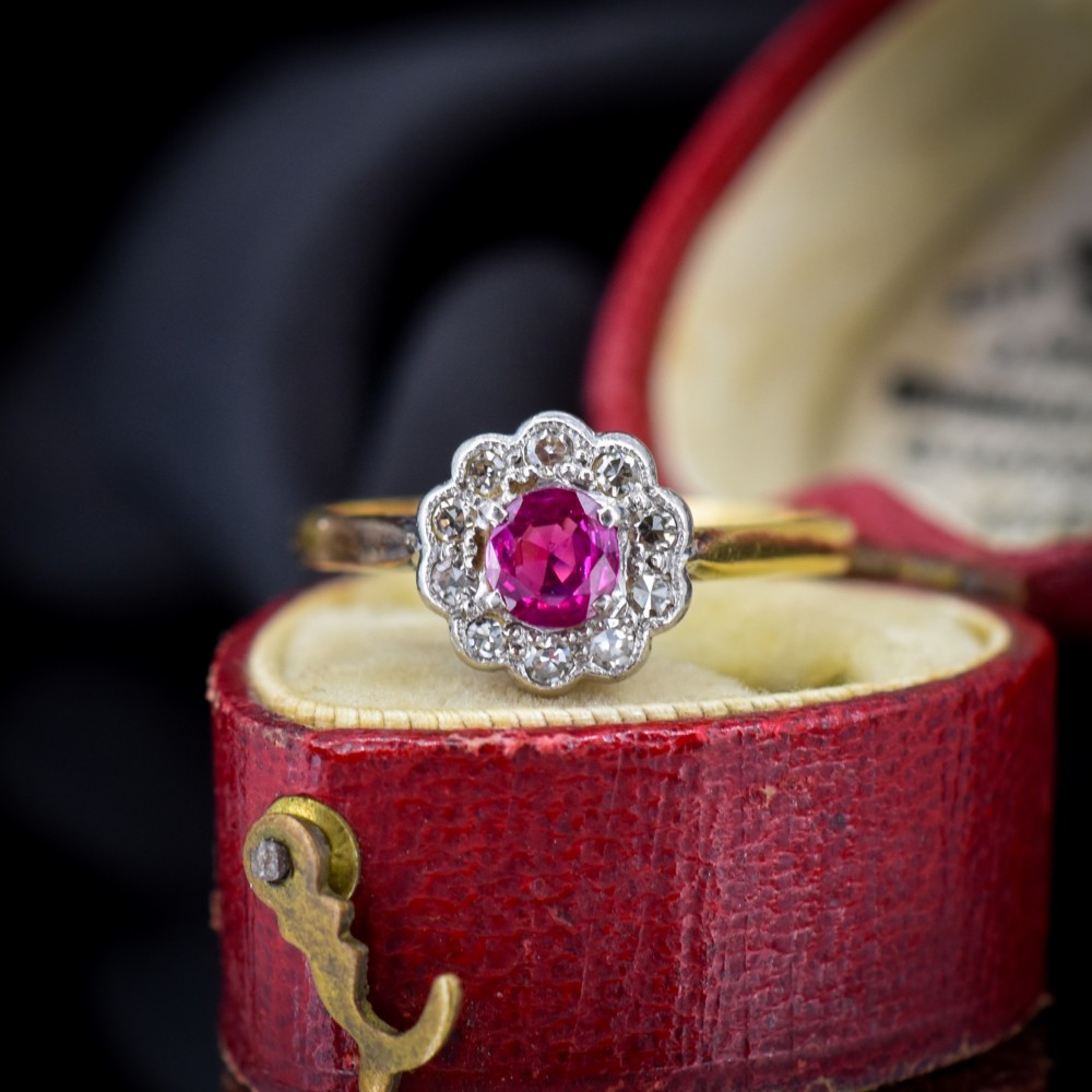 An Edwardian 18ct gold ring featuring a central oval faceted red ruby surrounded by a halo of small diamonds in a scalloped cluster setting. View IV