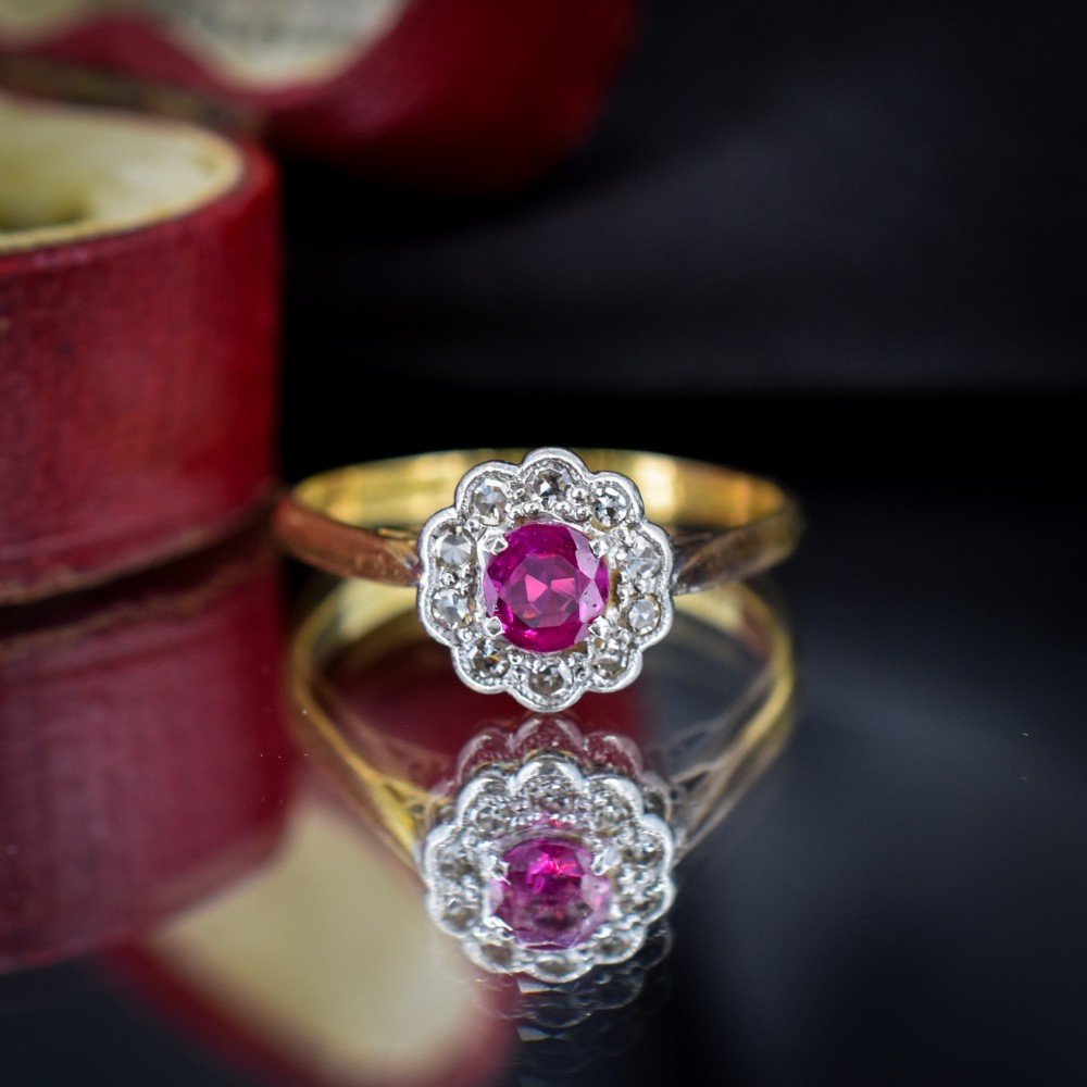 An Edwardian 18ct gold ring featuring a central oval faceted red ruby surrounded by a halo of small diamonds in a scalloped cluster setting. View V