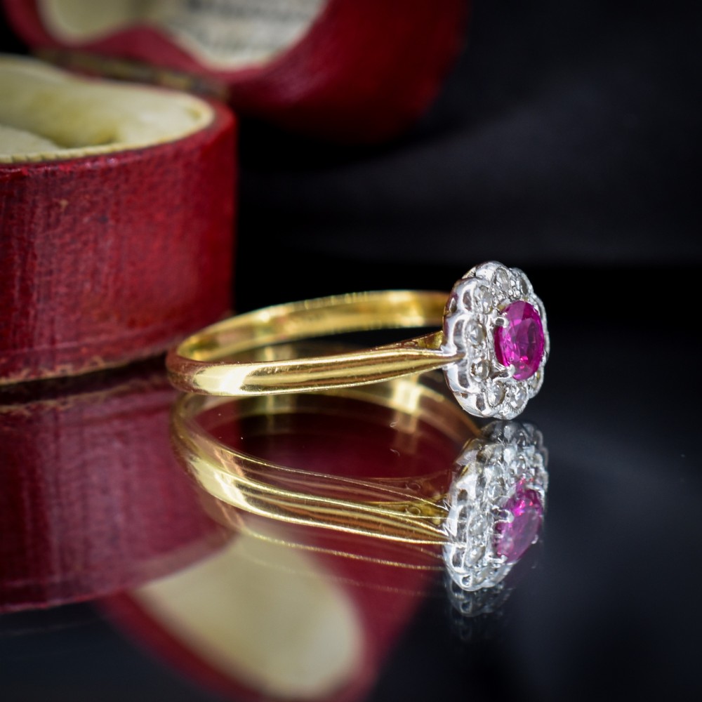 An Edwardian 18ct gold ring featuring a central oval faceted red ruby surrounded by a halo of small diamonds in a scalloped cluster setting. View VI