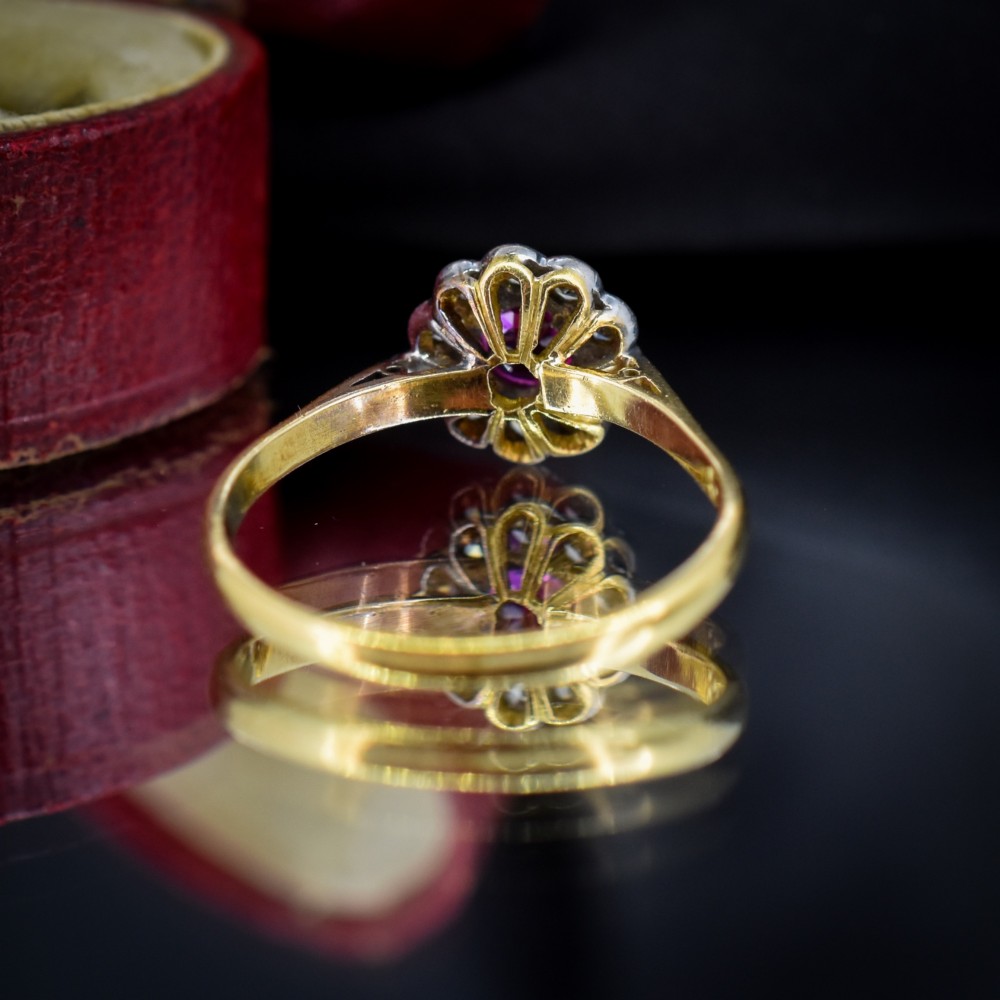An Edwardian 18ct gold ring featuring a central oval faceted red ruby surrounded by a halo of small diamonds in a scalloped cluster setting. View VII