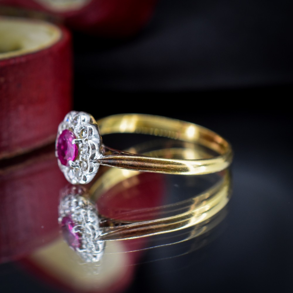 An Edwardian 18ct gold ring featuring a central oval faceted red ruby surrounded by a halo of small diamonds in a scalloped cluster setting. View VIII