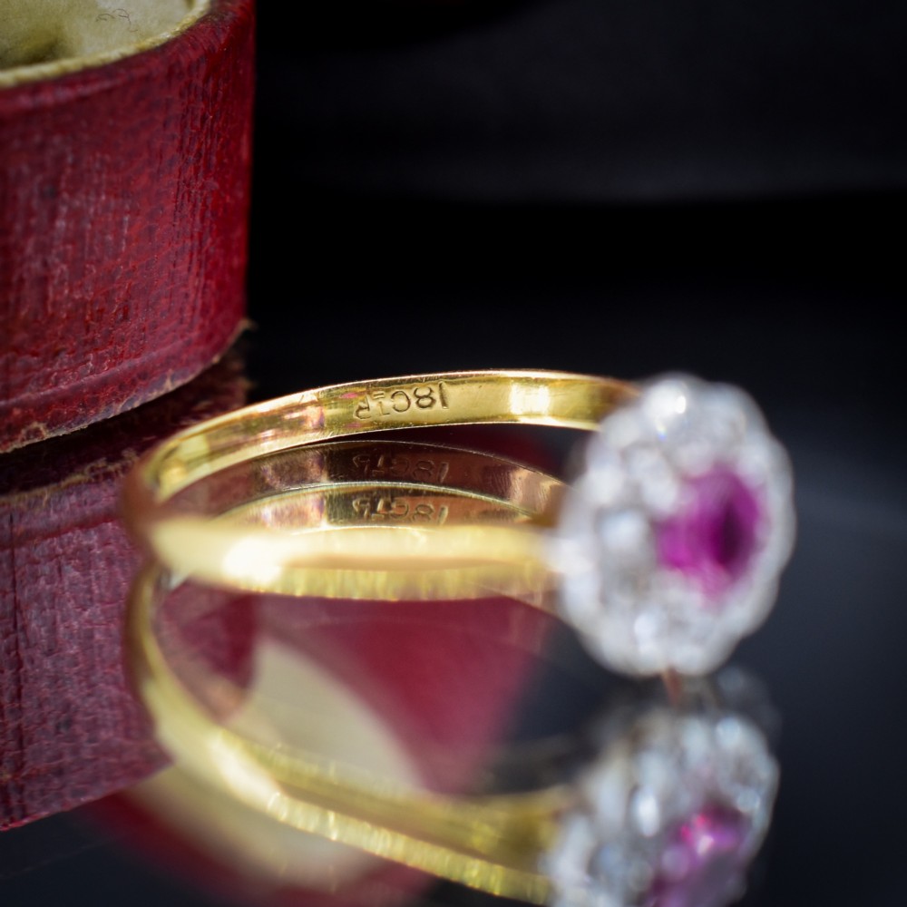 An Edwardian 18ct gold ring featuring a central oval faceted red ruby surrounded by a halo of small diamonds in a scalloped cluster setting. View IX