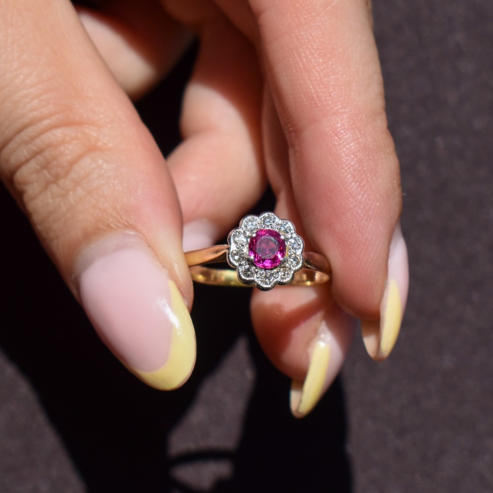 An Edwardian 18ct gold ring featuring a central oval faceted red ruby surrounded by a halo of small diamonds in a scalloped cluster setting. View X