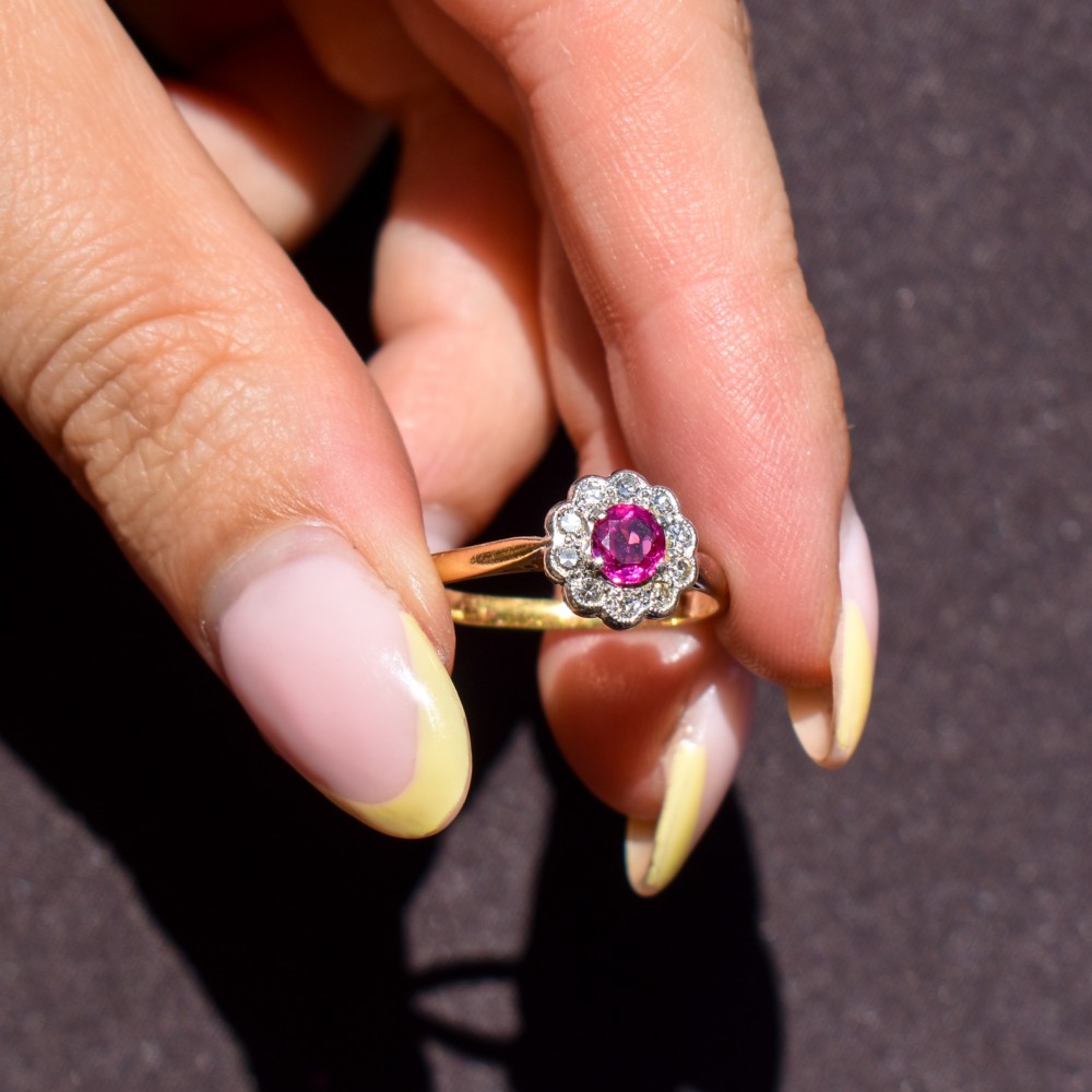 An Edwardian 18ct gold ring featuring a central oval faceted red ruby surrounded by a halo of small diamonds in a scalloped cluster setting. View XI