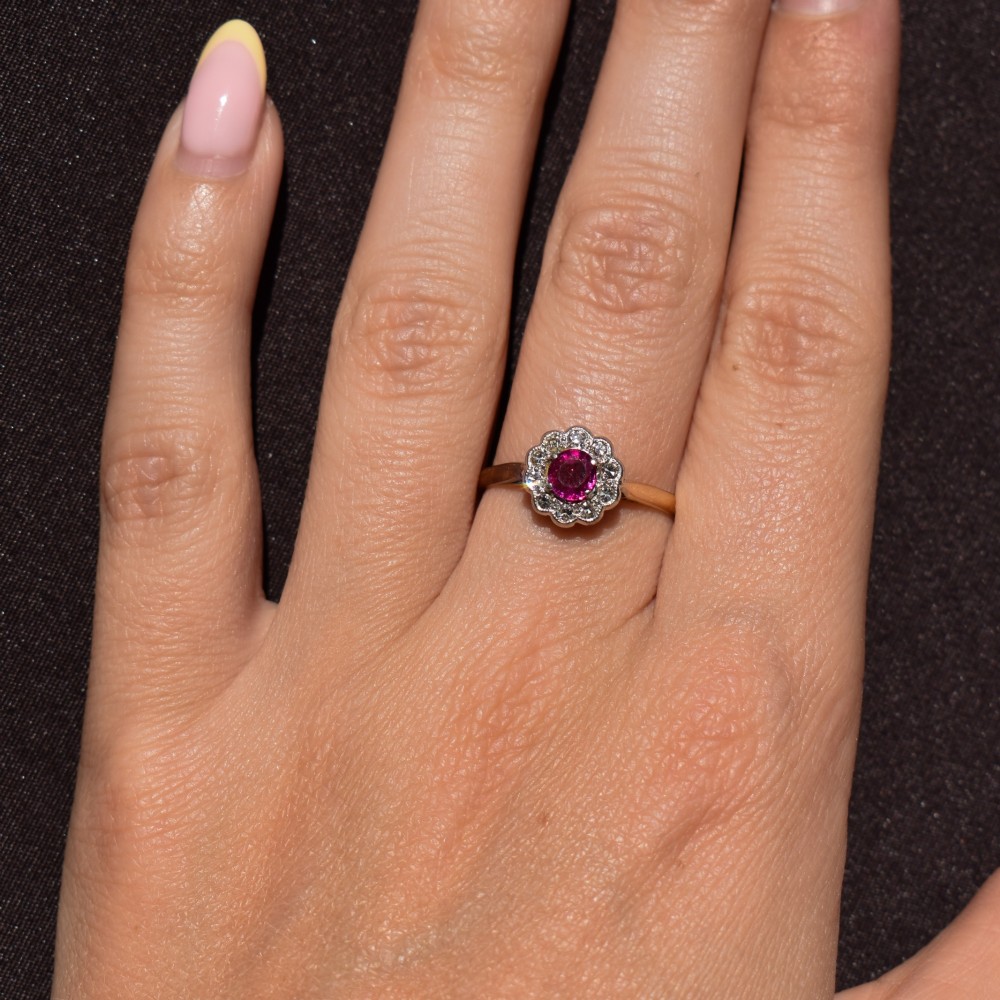 An Edwardian 18ct gold ring featuring a central oval faceted red ruby surrounded by a halo of small diamonds in a scalloped cluster setting. View XII