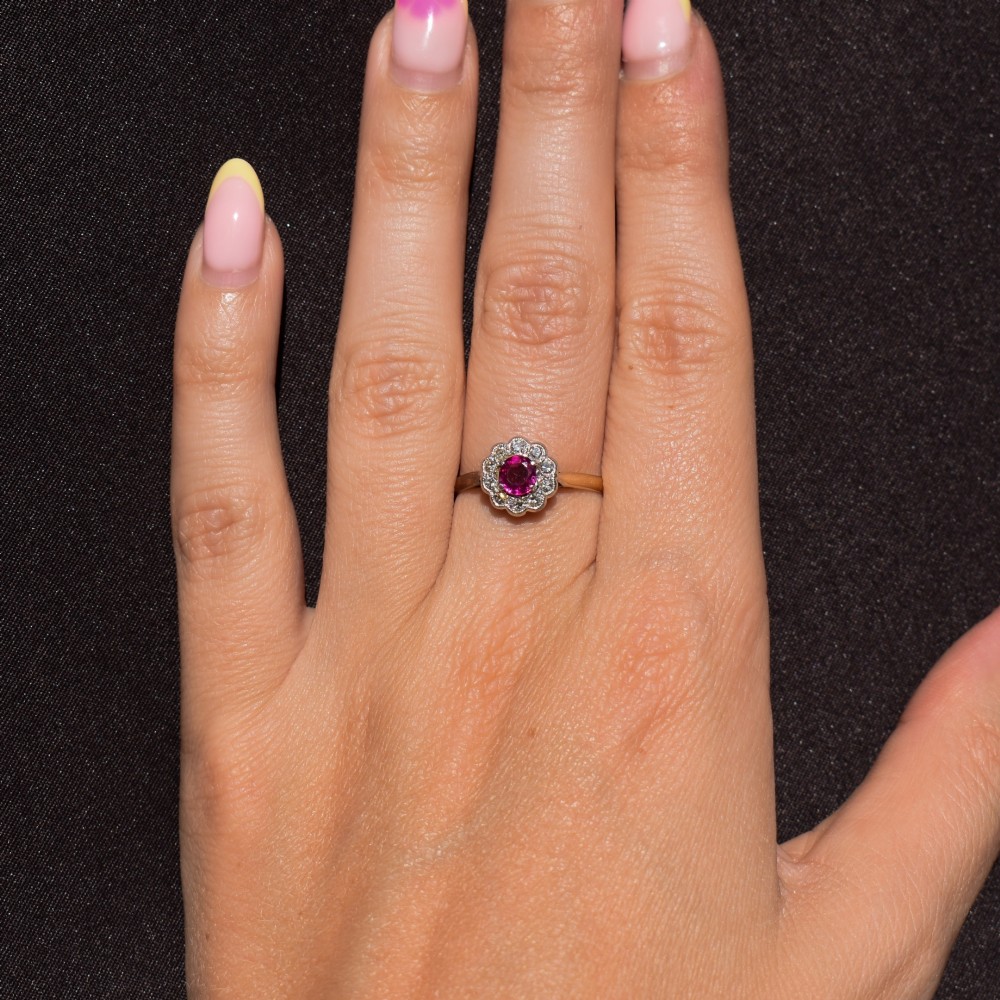 An Edwardian 18ct gold ring featuring a central oval faceted red ruby surrounded by a halo of small diamonds in a scalloped cluster setting. View XIII