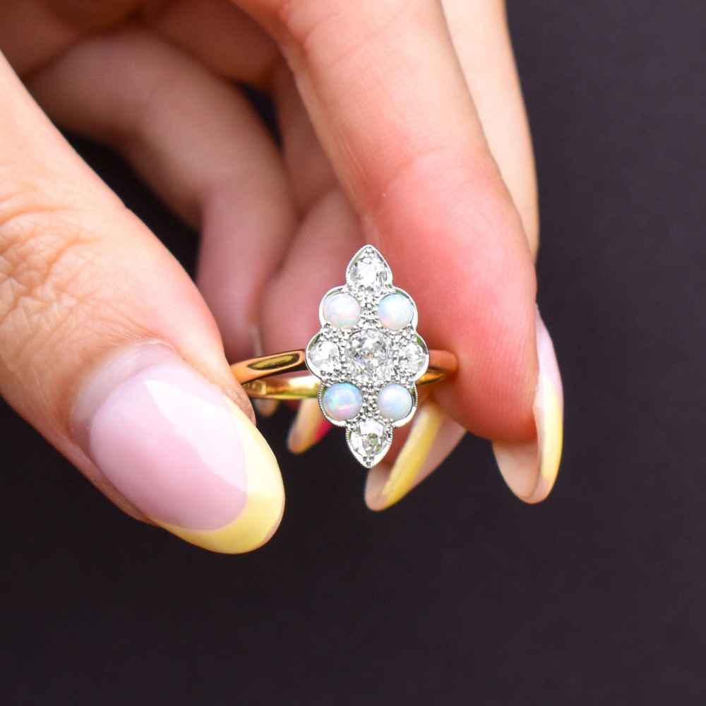 An Edwardian 18ct gold and platinum navette cluster ring featuring a central old cut diamond bordered by four cabochon opals and additional diamond accents. View X