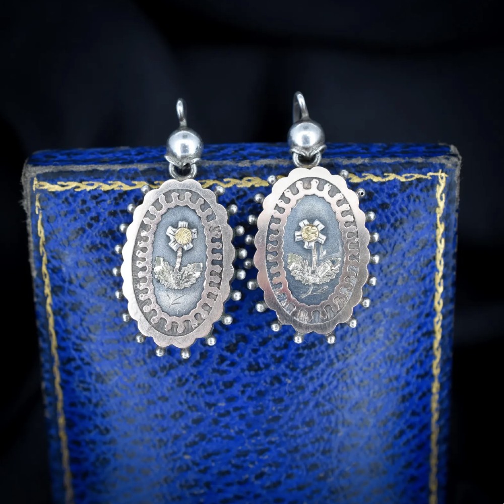 A pair of oval Victorian sterling silver drop earrings featuring central engraved flowers, scalloped inner borders, and beaded outer rims.