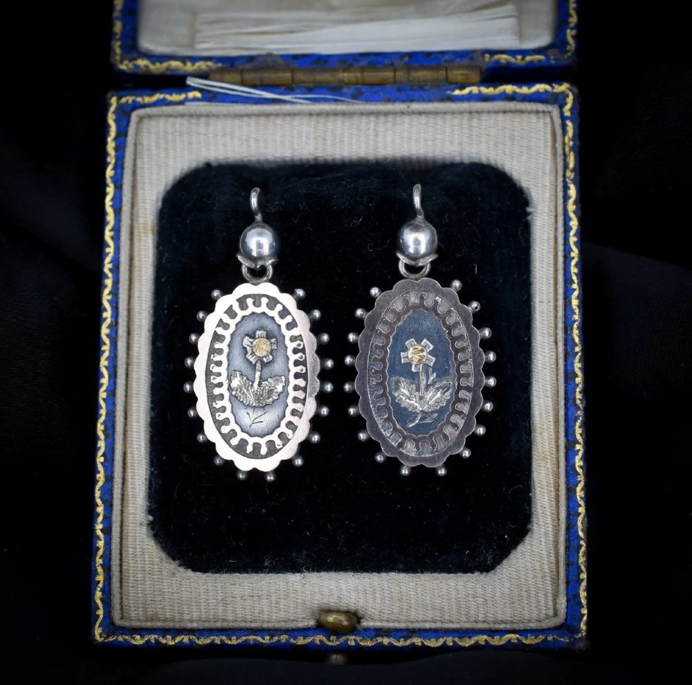 A pair of oval Victorian sterling silver drop earrings featuring central engraved flowers, scalloped inner borders, and beaded outer rims. View V