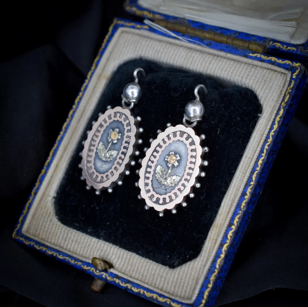 A pair of oval Victorian sterling silver drop earrings featuring central engraved flowers, scalloped inner borders, and beaded outer rims. View VI
