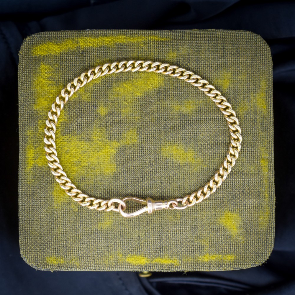 An antique 9ct yellow gold uniform curb link chain bracelet with a dog clip clasp arrangement resting on a textured olive display pad. View II