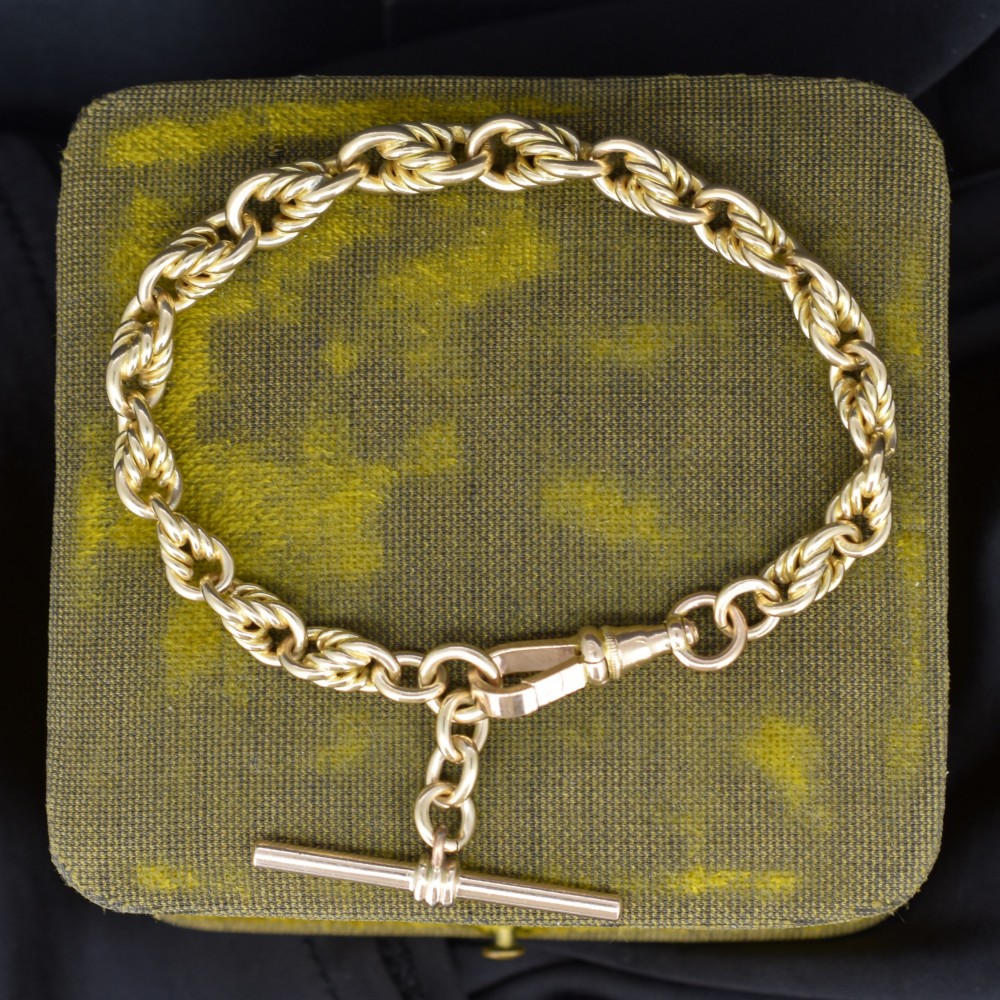 An Edwardian 9ct yellow gold love knot chain bracelet with a suspended T-bar and swivel clasp fastener on a textured green surface.