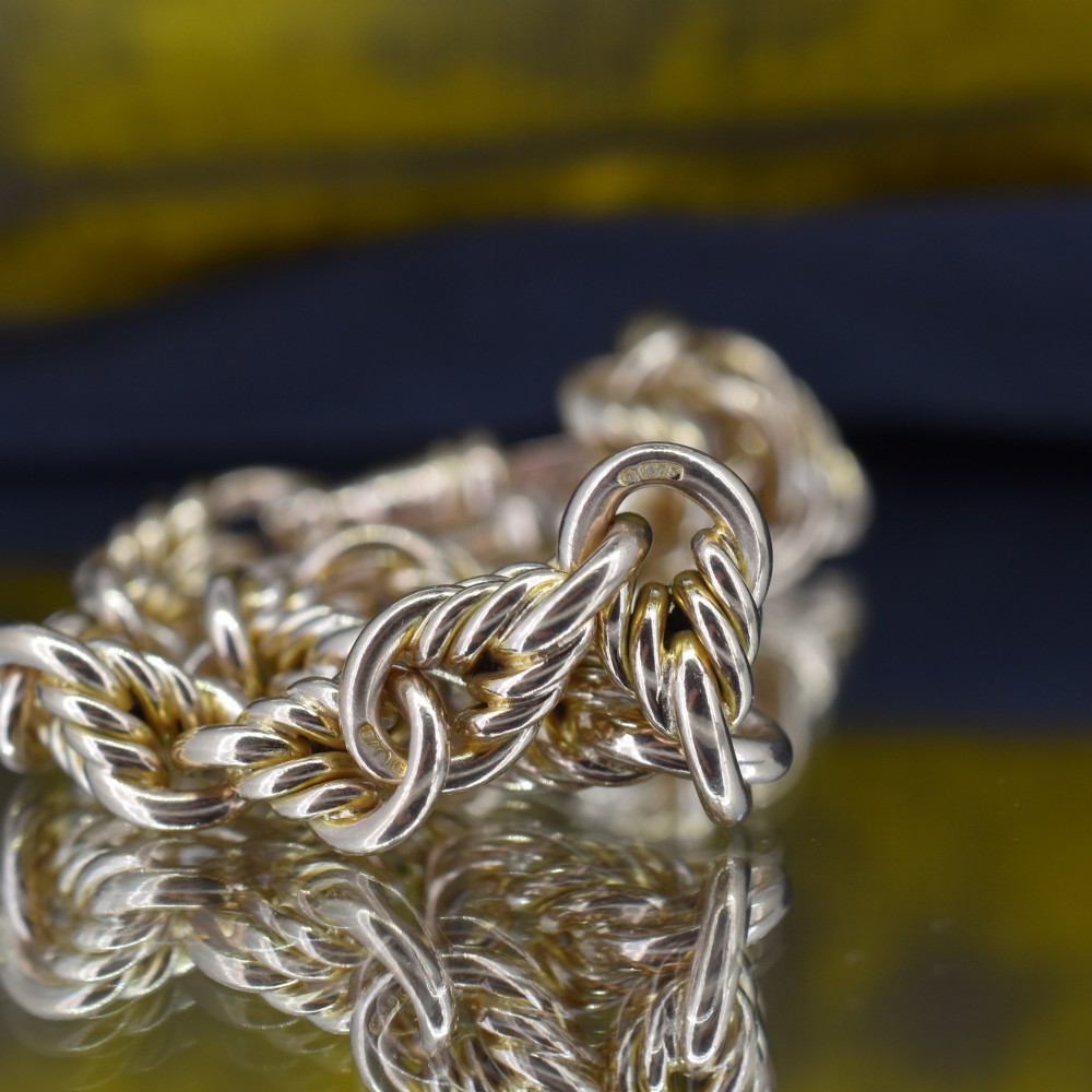 An Edwardian 9ct yellow gold love knot chain bracelet with a suspended T-bar and swivel clasp fastener on a textured green surface. View V