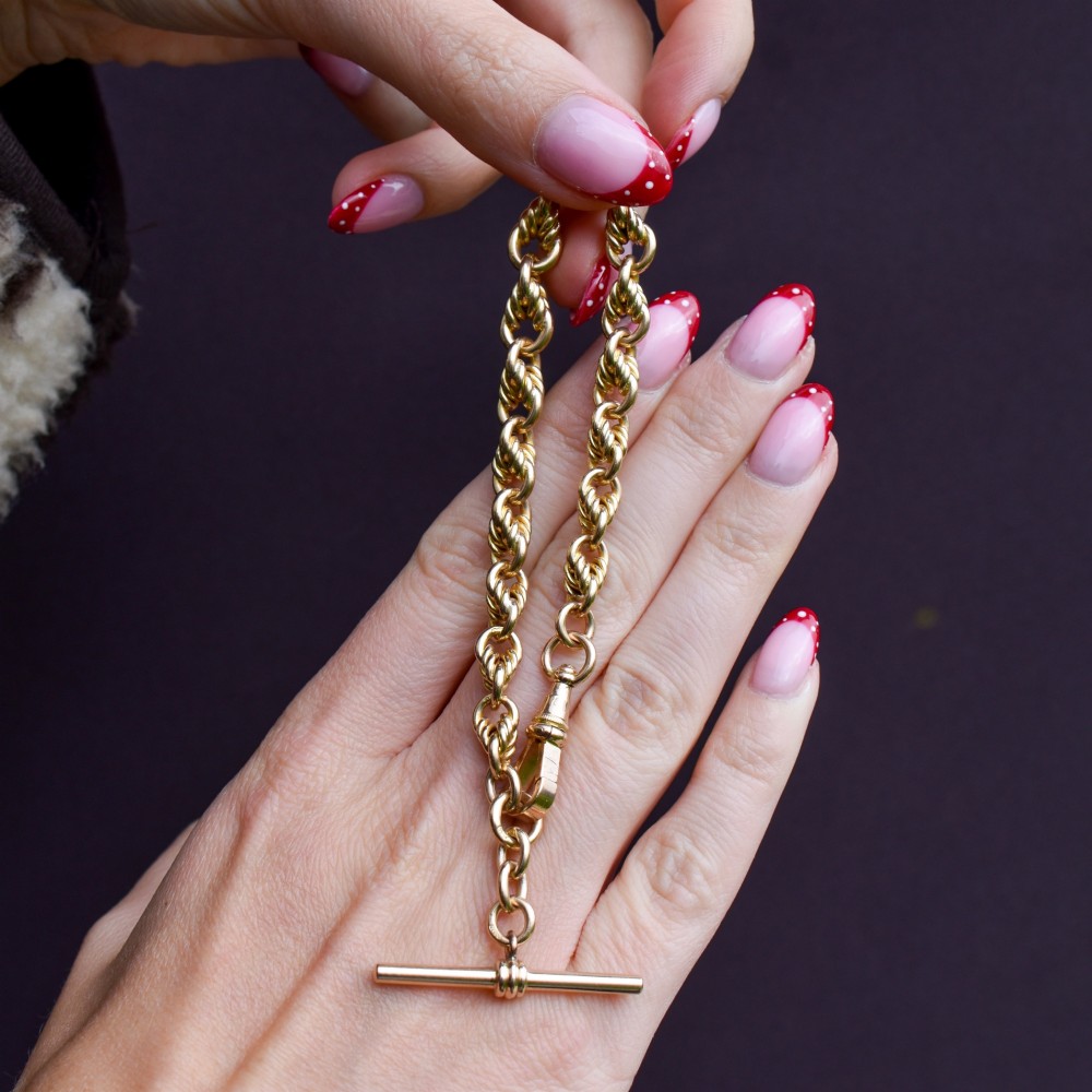 An Edwardian 9ct yellow gold love knot chain bracelet with a suspended T-bar and swivel clasp fastener on a textured green surface. View VII