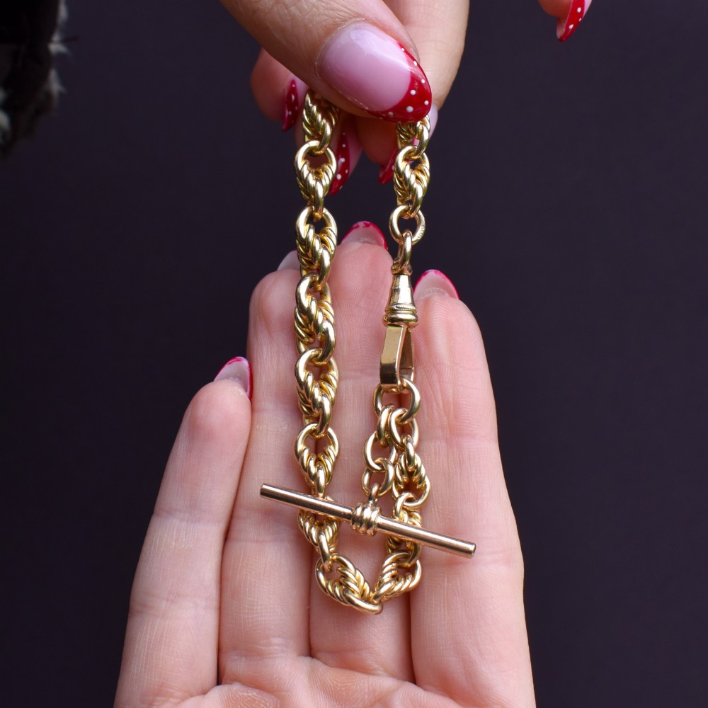 An Edwardian 9ct yellow gold love knot chain bracelet with a suspended T-bar and swivel clasp fastener on a textured green surface. View IX