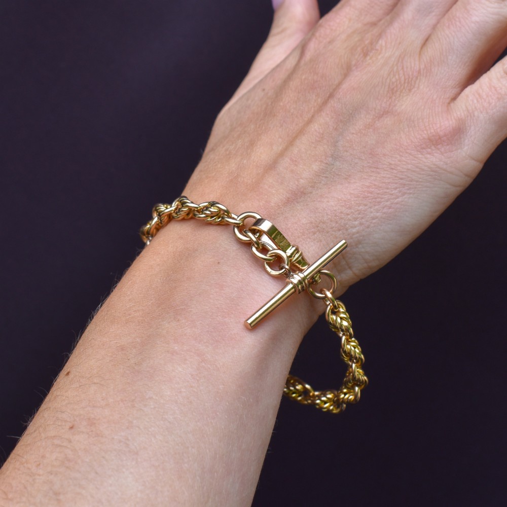 An Edwardian 9ct yellow gold love knot chain bracelet with a suspended T-bar and swivel clasp fastener on a textured green surface. View X