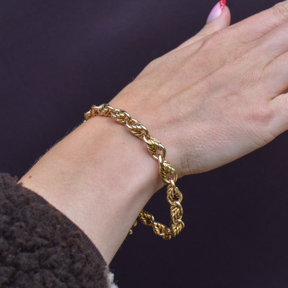 An Edwardian 9ct yellow gold love knot chain bracelet with a suspended T-bar and swivel clasp fastener on a textured green surface. View XI