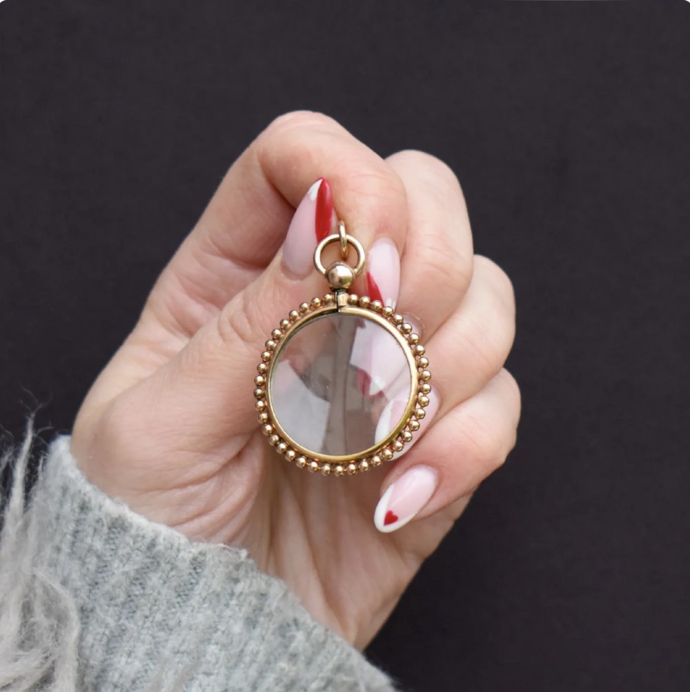 A circular antique gold locket featuring clear glass panels surrounded by a beaded metal border and attached to a suspension loop. View III