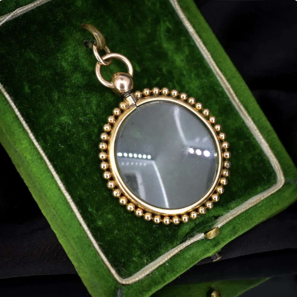 A circular antique gold locket featuring clear glass panels surrounded by a beaded metal border and attached to a suspension loop. View IV