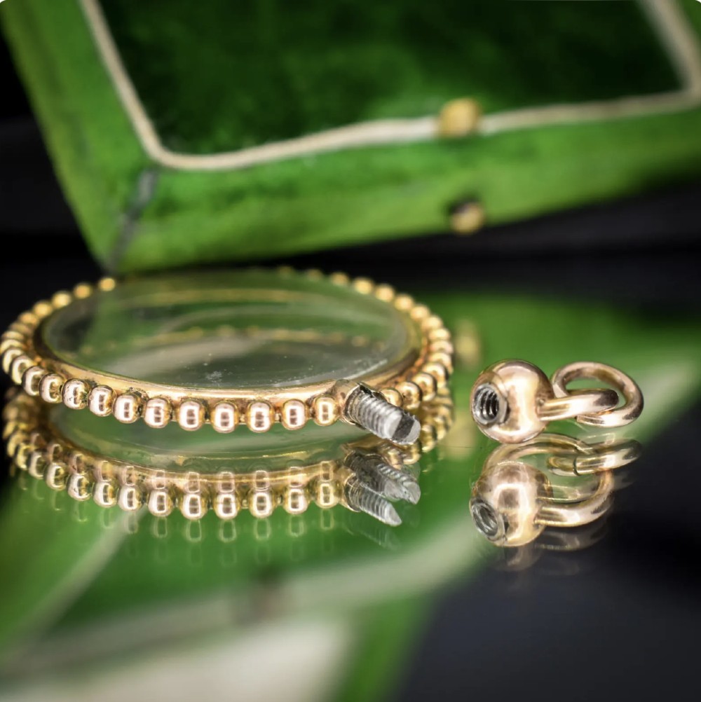 A circular antique gold locket featuring clear glass panels surrounded by a beaded metal border and attached to a suspension loop. View VI