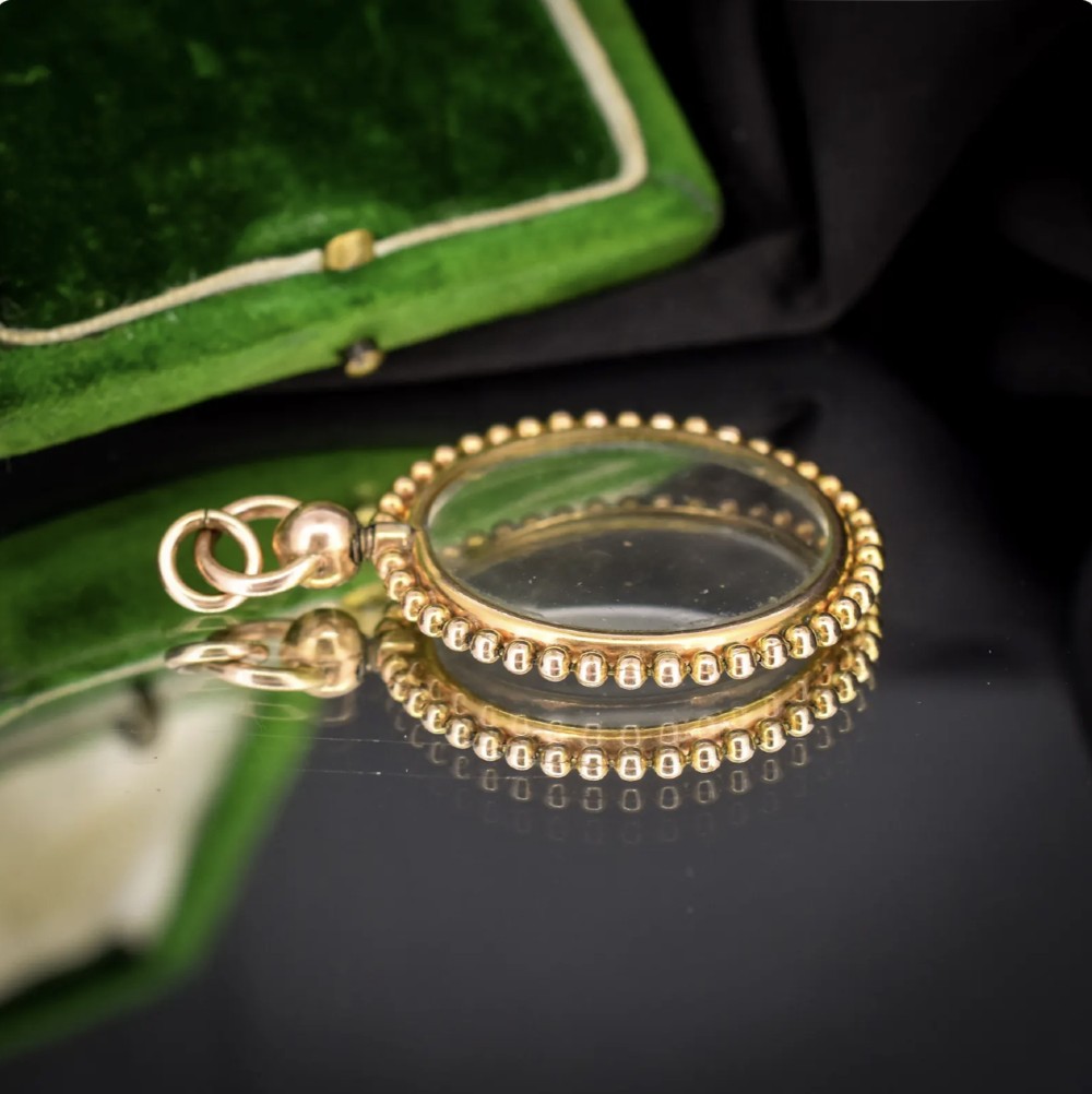 A circular antique gold locket featuring clear glass panels surrounded by a beaded metal border and attached to a suspension loop. View VII
