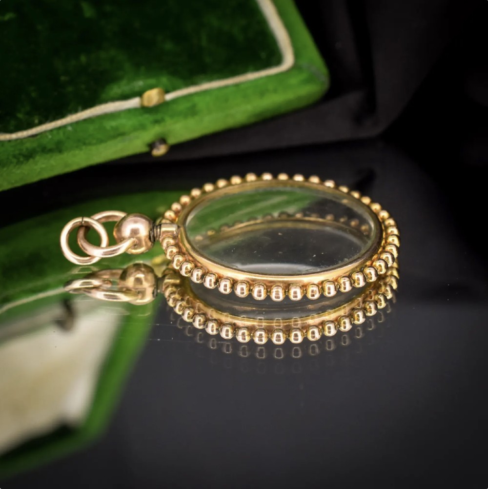 A circular antique gold locket featuring clear glass panels surrounded by a beaded metal border and attached to a suspension loop. View IX