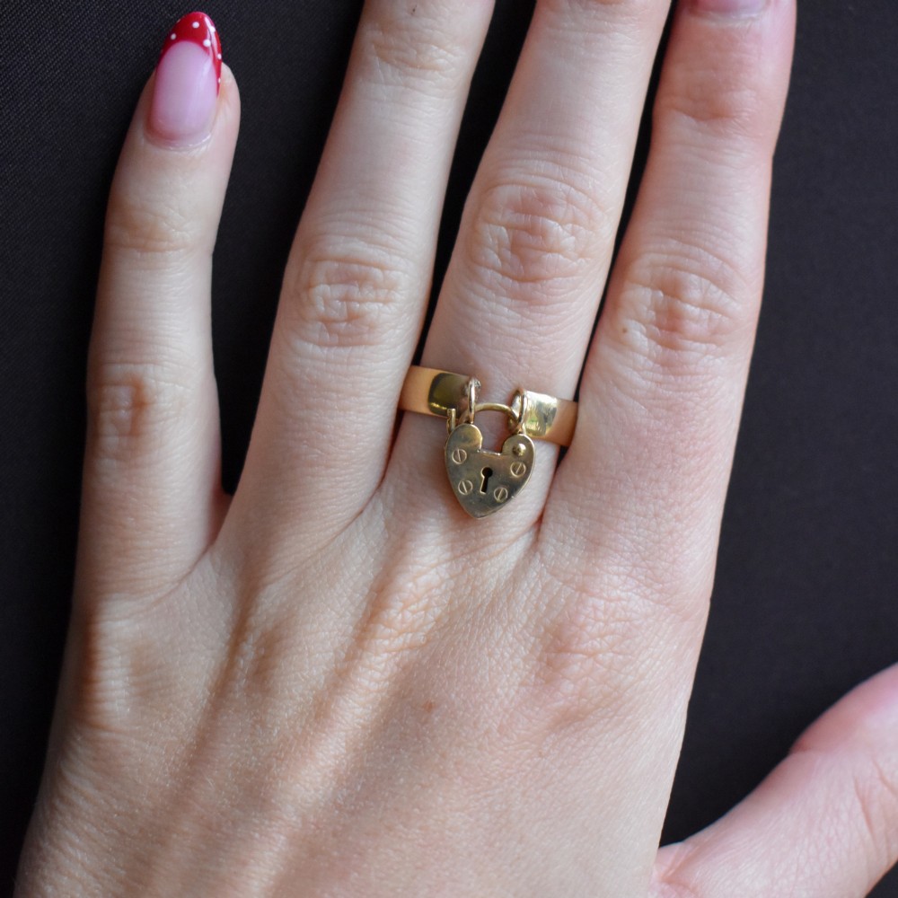 A late Victorian 9ct yellow gold band ring featuring a suspended heart-shaped padlock charm with keyhole detail. View X