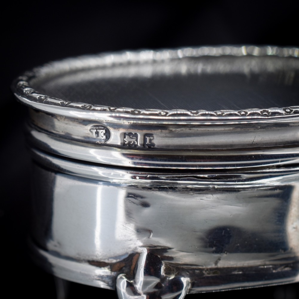 An oval silver trinket box resting on three cabriole legs with a patterned rim along the lid. View II