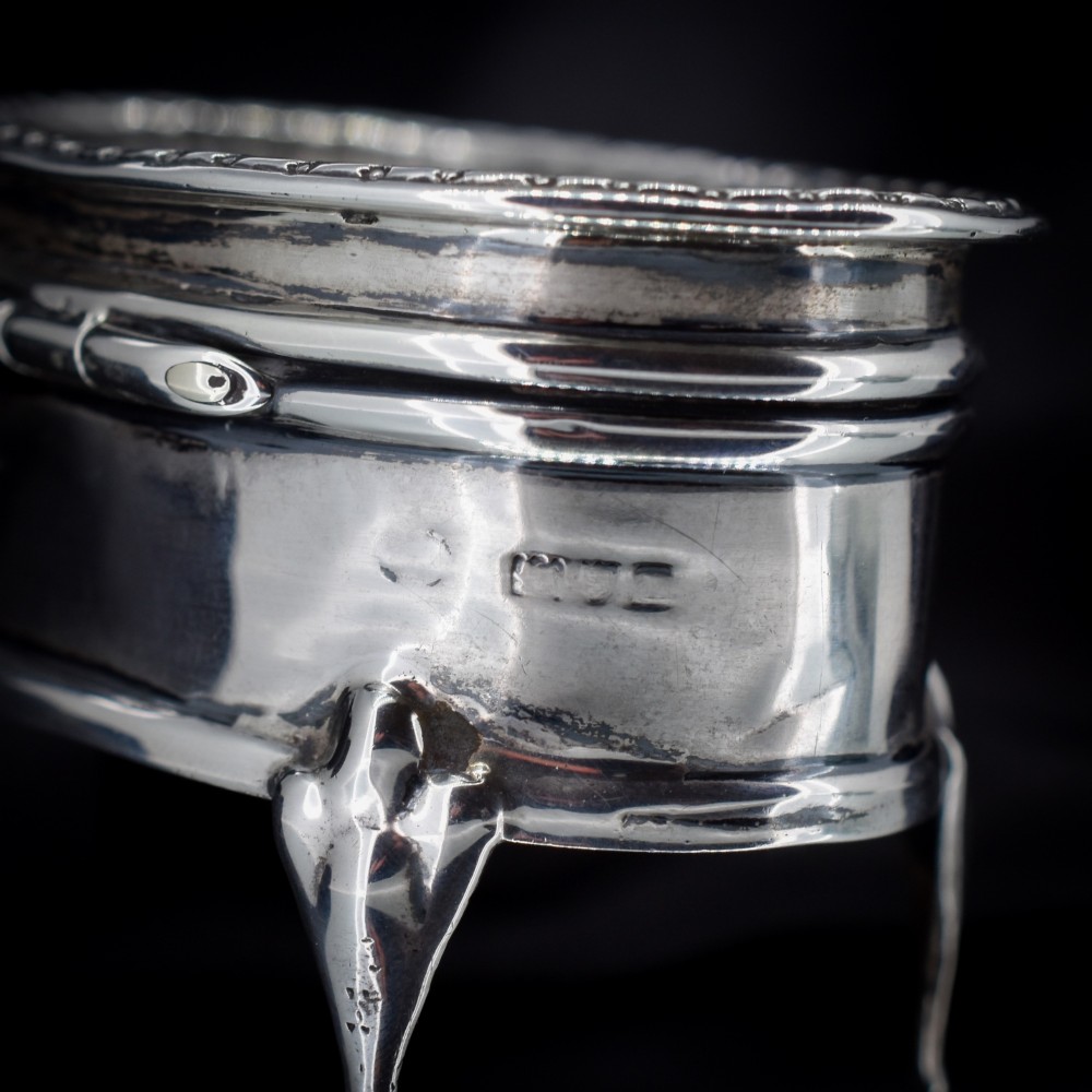 An oval silver trinket box resting on three cabriole legs with a patterned rim along the lid. View V
