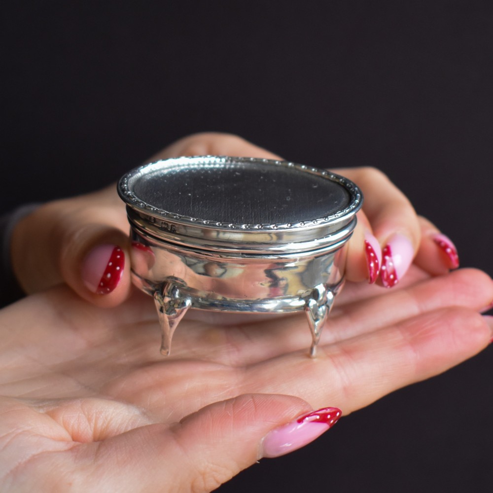 An oval silver trinket box resting on three cabriole legs with a patterned rim along the lid.