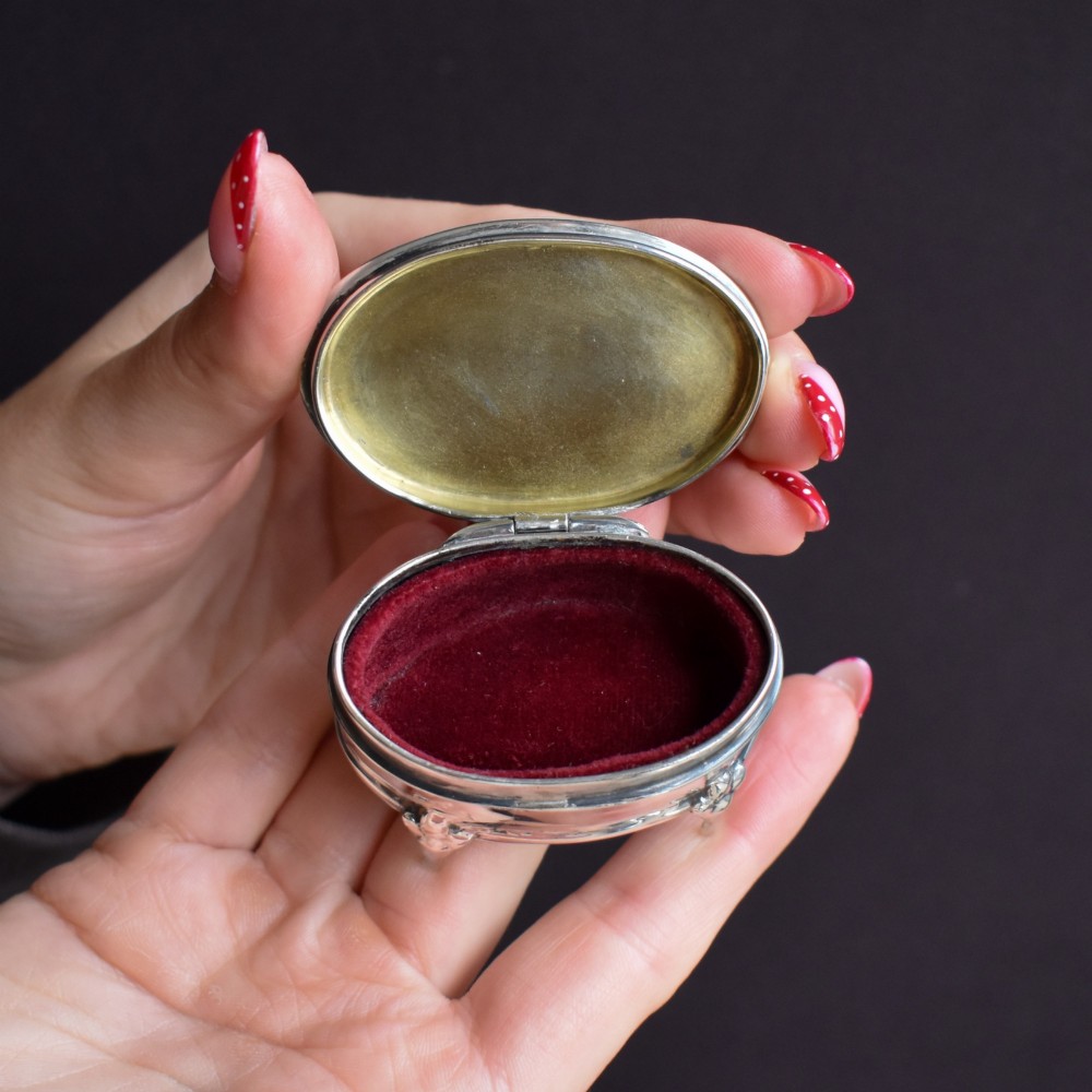 An oval silver trinket box resting on three cabriole legs with a patterned rim along the lid. View VII