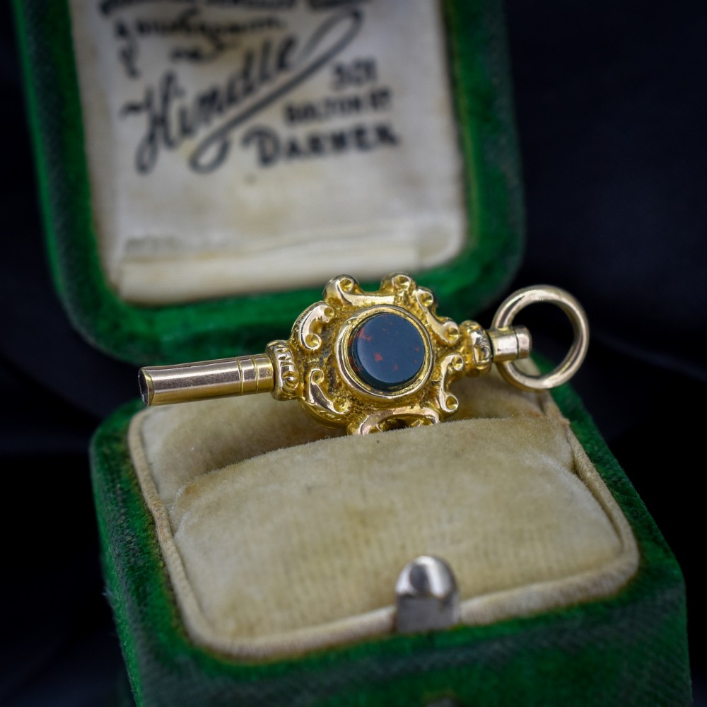 An antique gold watch key pendant featuring scrollwork ornamentation around a central dark green bloodstone seal, topped with a suspension ring.