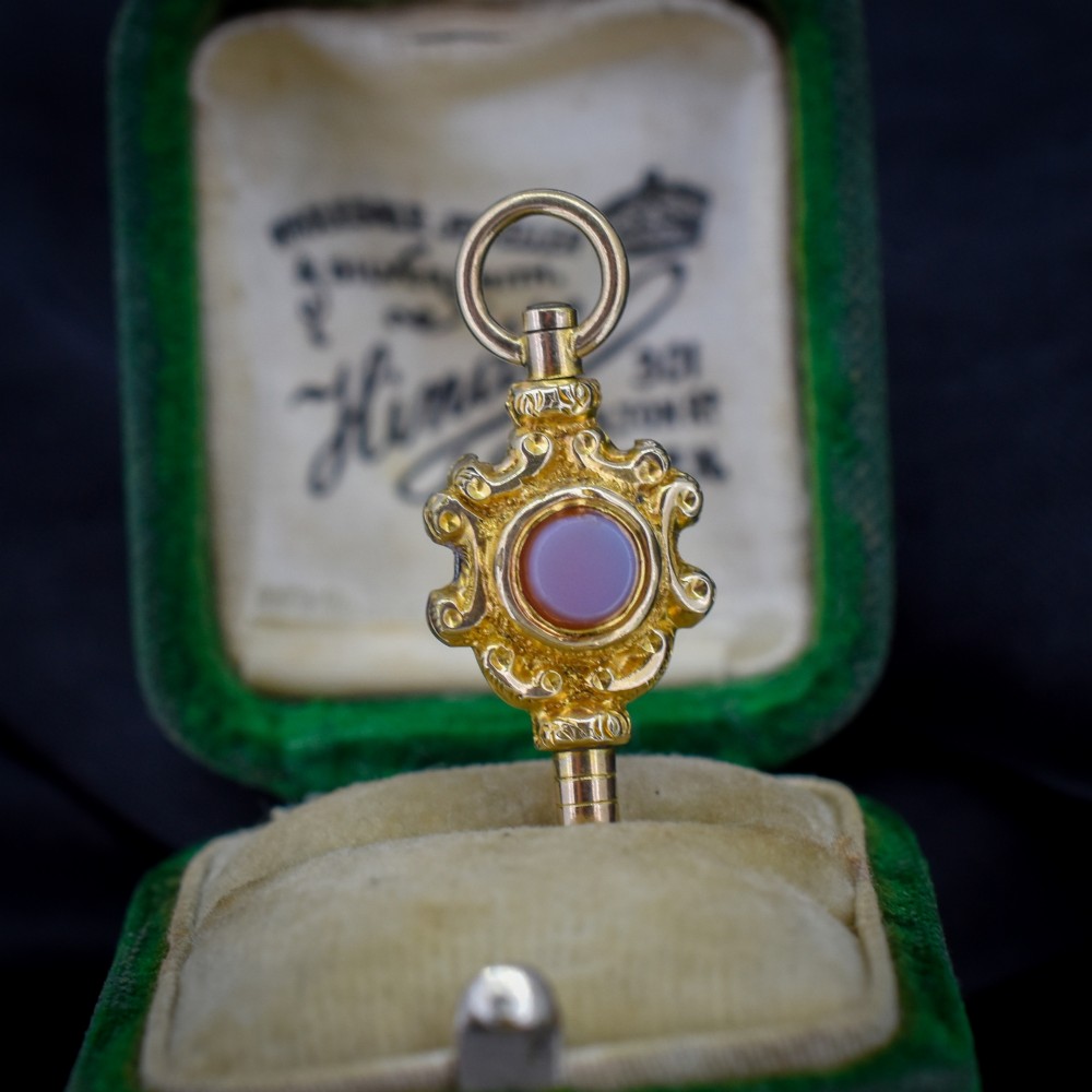 An antique gold watch key pendant featuring scrollwork ornamentation around a central dark green bloodstone seal, topped with a suspension ring. View III
