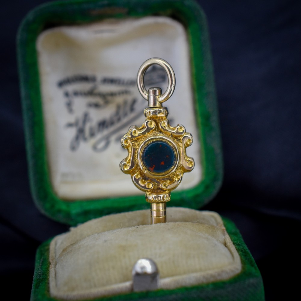 An antique gold watch key pendant featuring scrollwork ornamentation around a central dark green bloodstone seal, topped with a suspension ring. View IV
