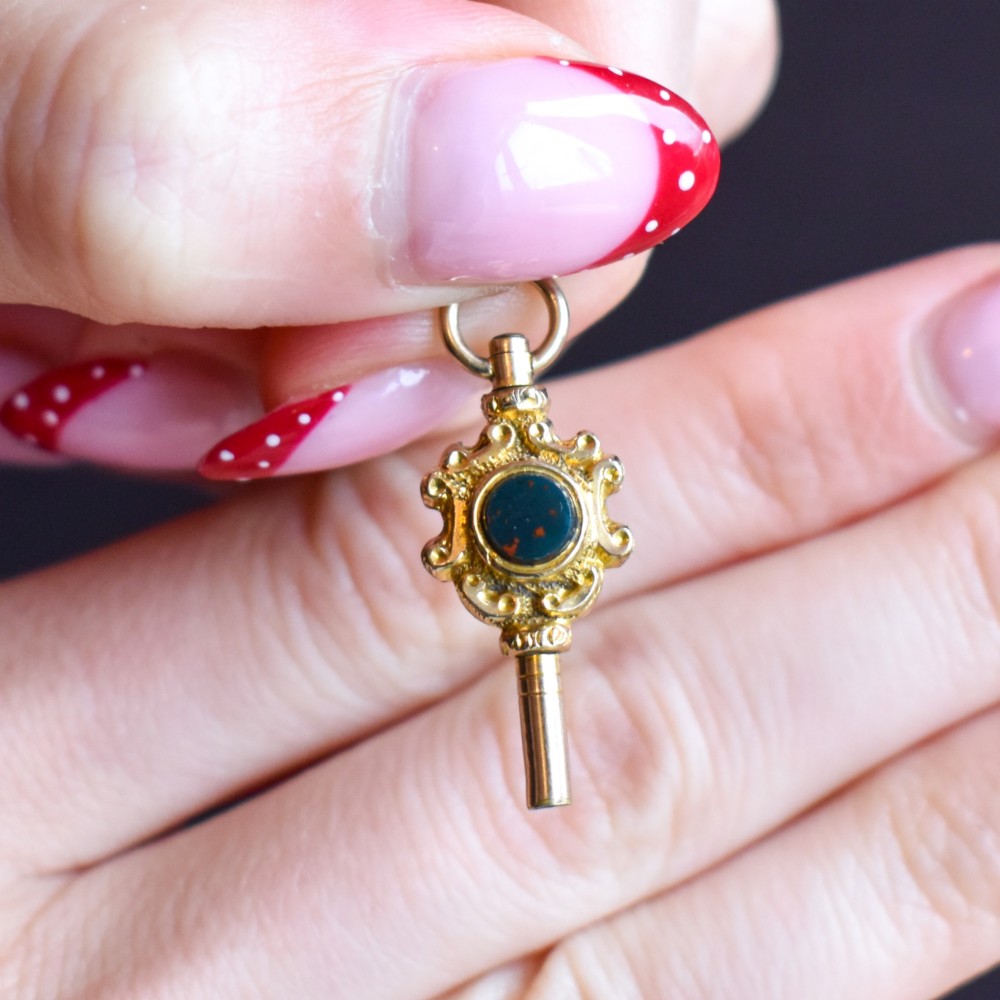An antique gold watch key pendant featuring scrollwork ornamentation around a central dark green bloodstone seal, topped with a suspension ring. View VII