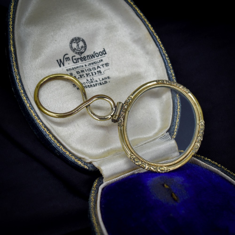 A Georgian rolled gold quizzing glass pendant featuring an engraved circular bezel and an S-scroll handle, resting inside a velvet-lined case.