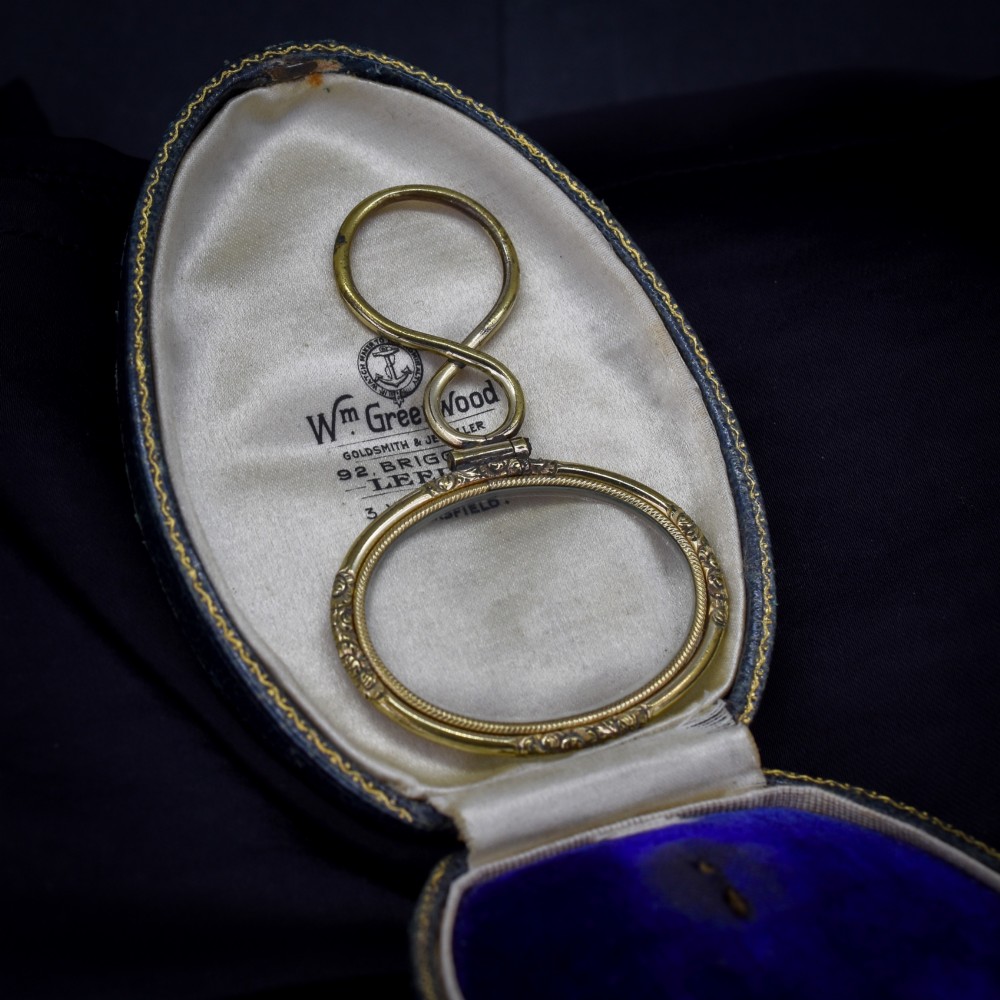 A Georgian rolled gold quizzing glass pendant featuring an engraved circular bezel and an S-scroll handle, resting inside a velvet-lined case. View IV