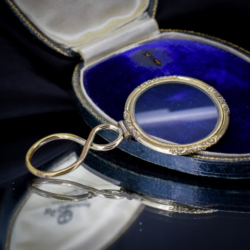 A Georgian rolled gold quizzing glass pendant featuring an engraved circular bezel and an S-scroll handle, resting inside a velvet-lined case. View V