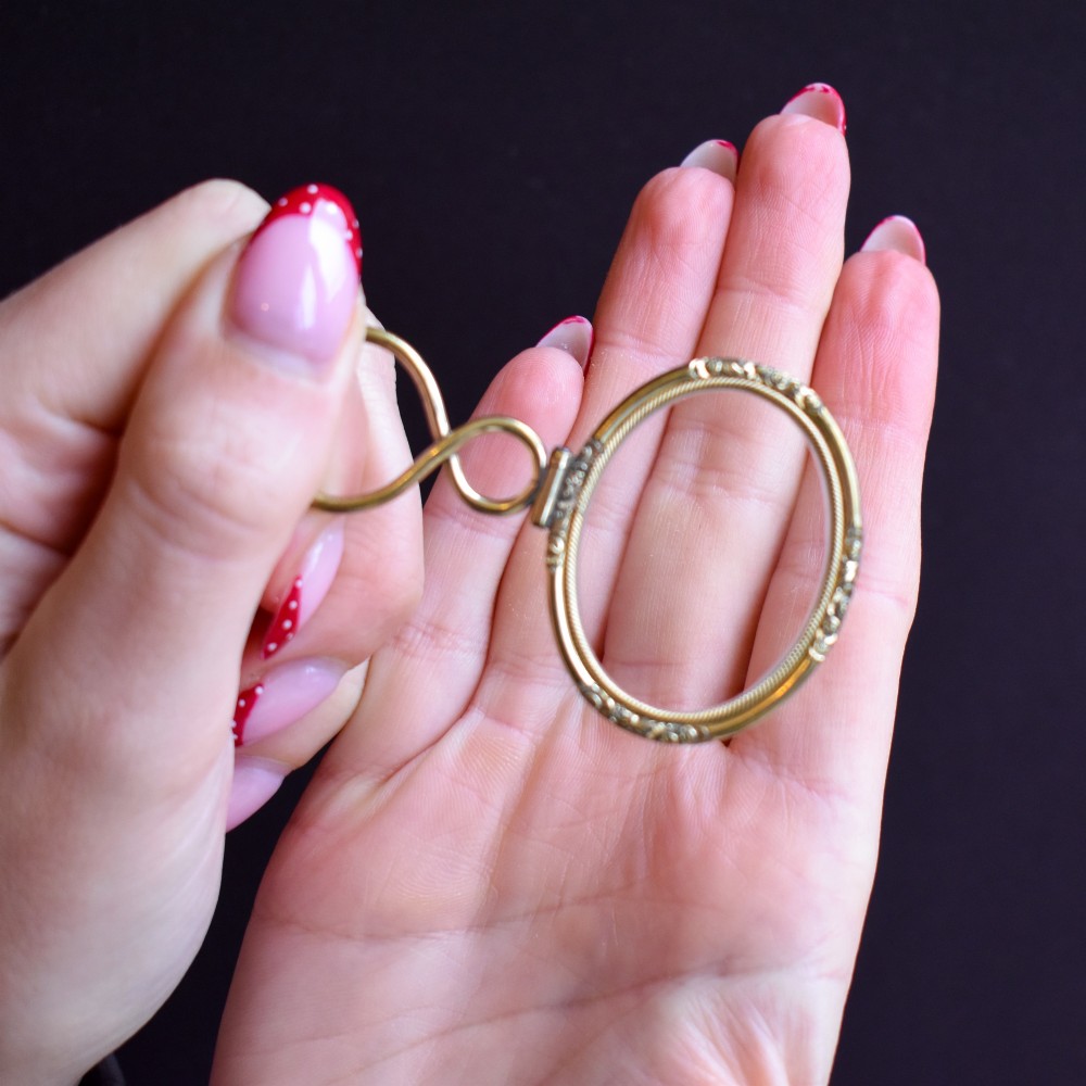 A Georgian rolled gold quizzing glass pendant featuring an engraved circular bezel and an S-scroll handle, resting inside a velvet-lined case. View VI