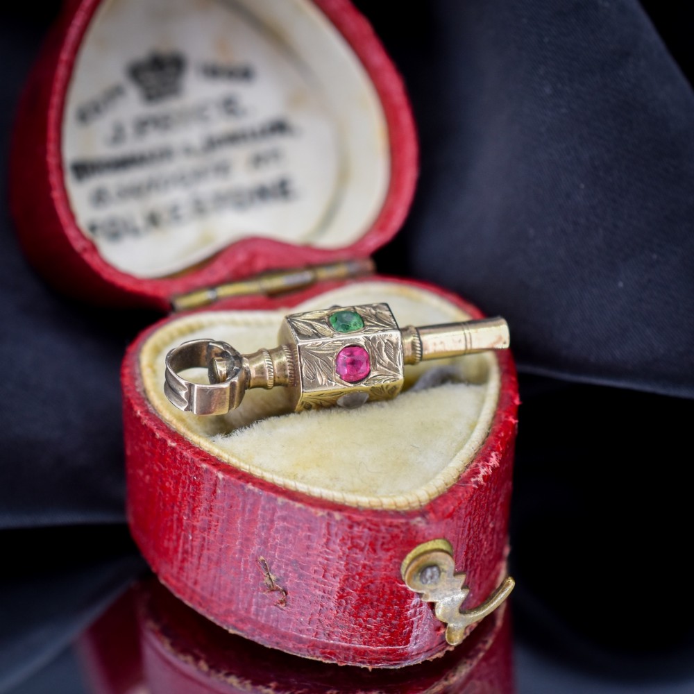 An antique yellow gold barrel watch key fob pendant with engraved scrollwork and a vertical setting of acrostic gemstones.