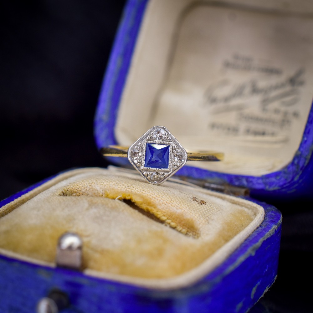 An Art Deco ring displaying a square blue sapphire surrounded by diamonds in a diamond-shaped geometric setting on an 18ct yellow gold band. View II