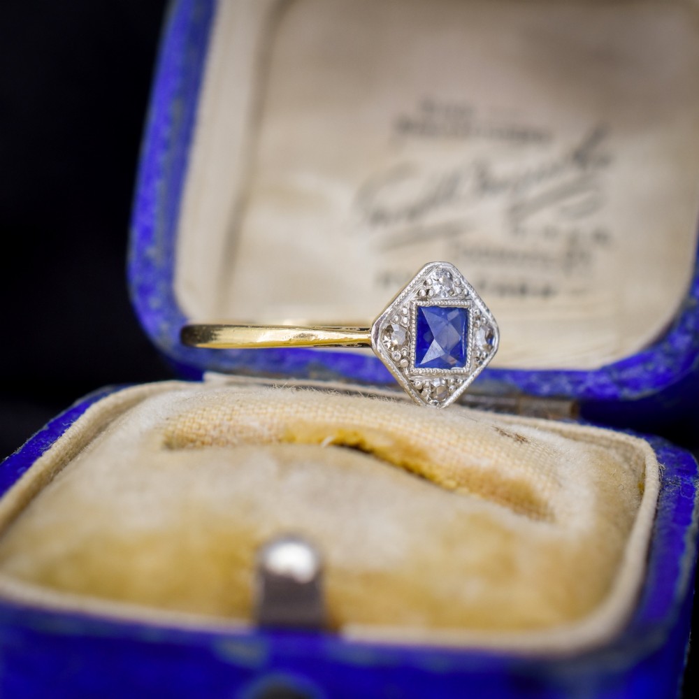 An Art Deco ring displaying a square blue sapphire surrounded by diamonds in a diamond-shaped geometric setting on an 18ct yellow gold band. View III