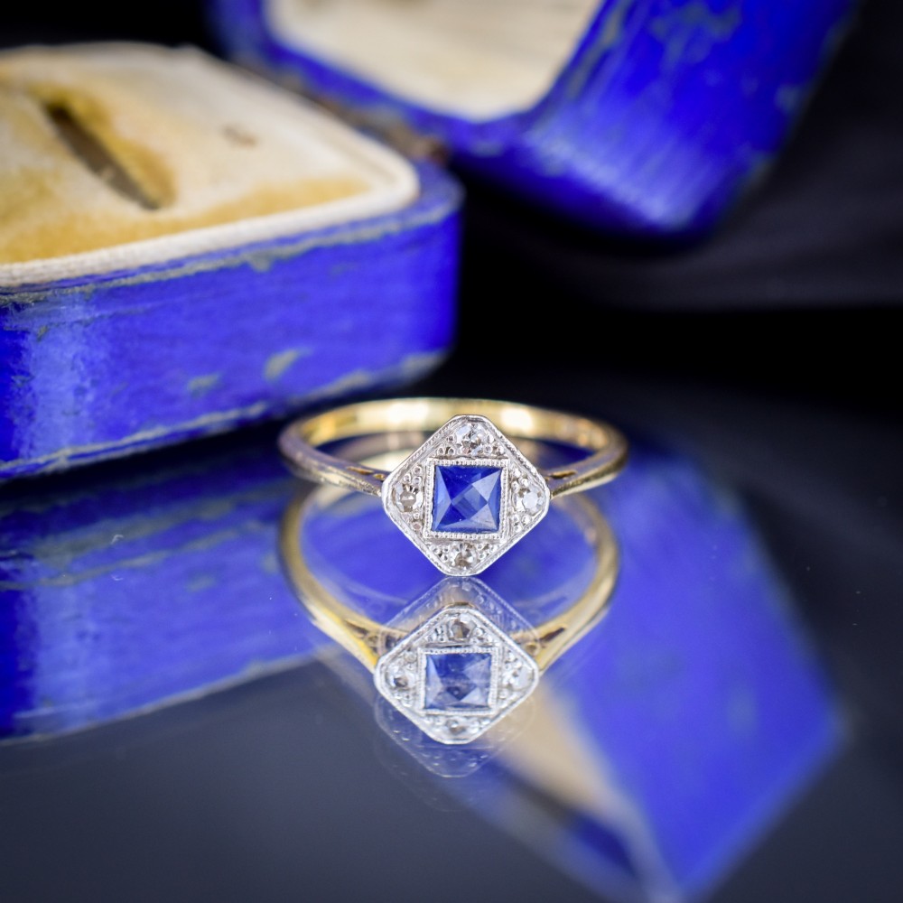 An Art Deco ring displaying a square blue sapphire surrounded by diamonds in a diamond-shaped geometric setting on an 18ct yellow gold band. View IV