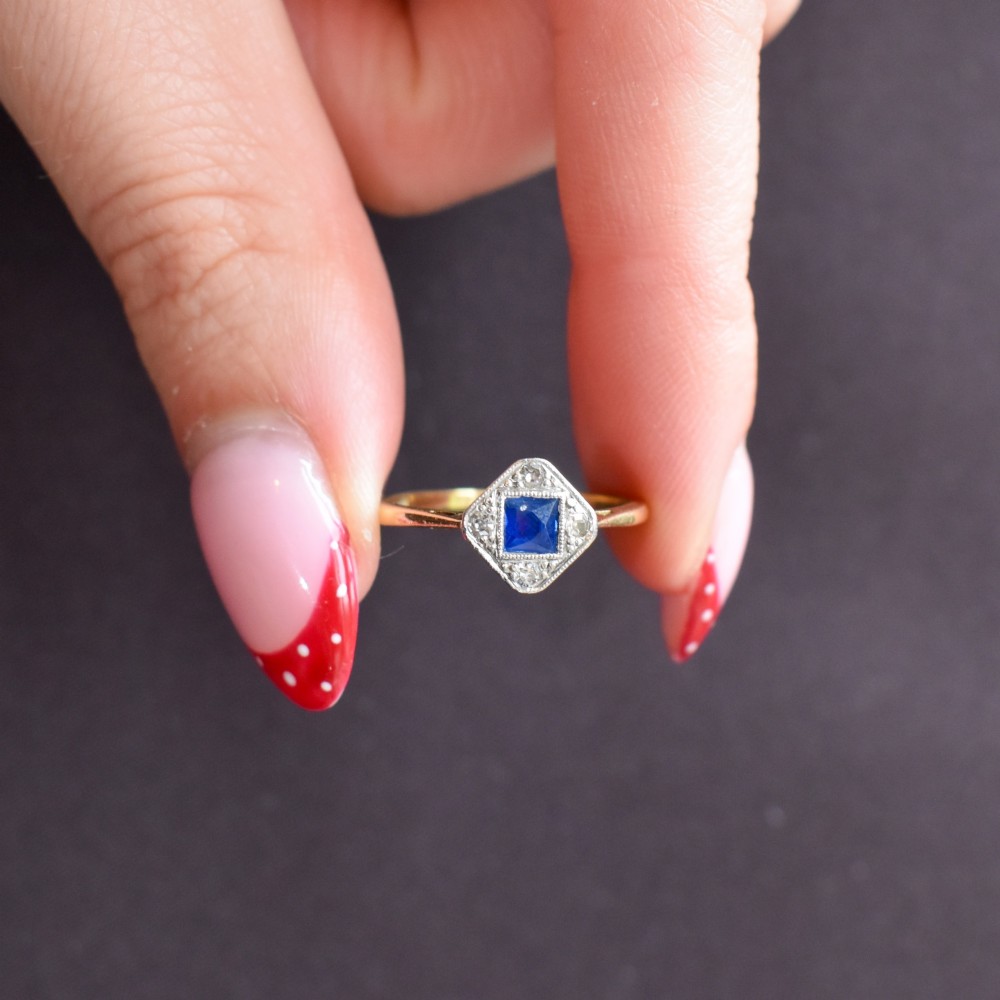 An Art Deco ring displaying a square blue sapphire surrounded by diamonds in a diamond-shaped geometric setting on an 18ct yellow gold band. View IX