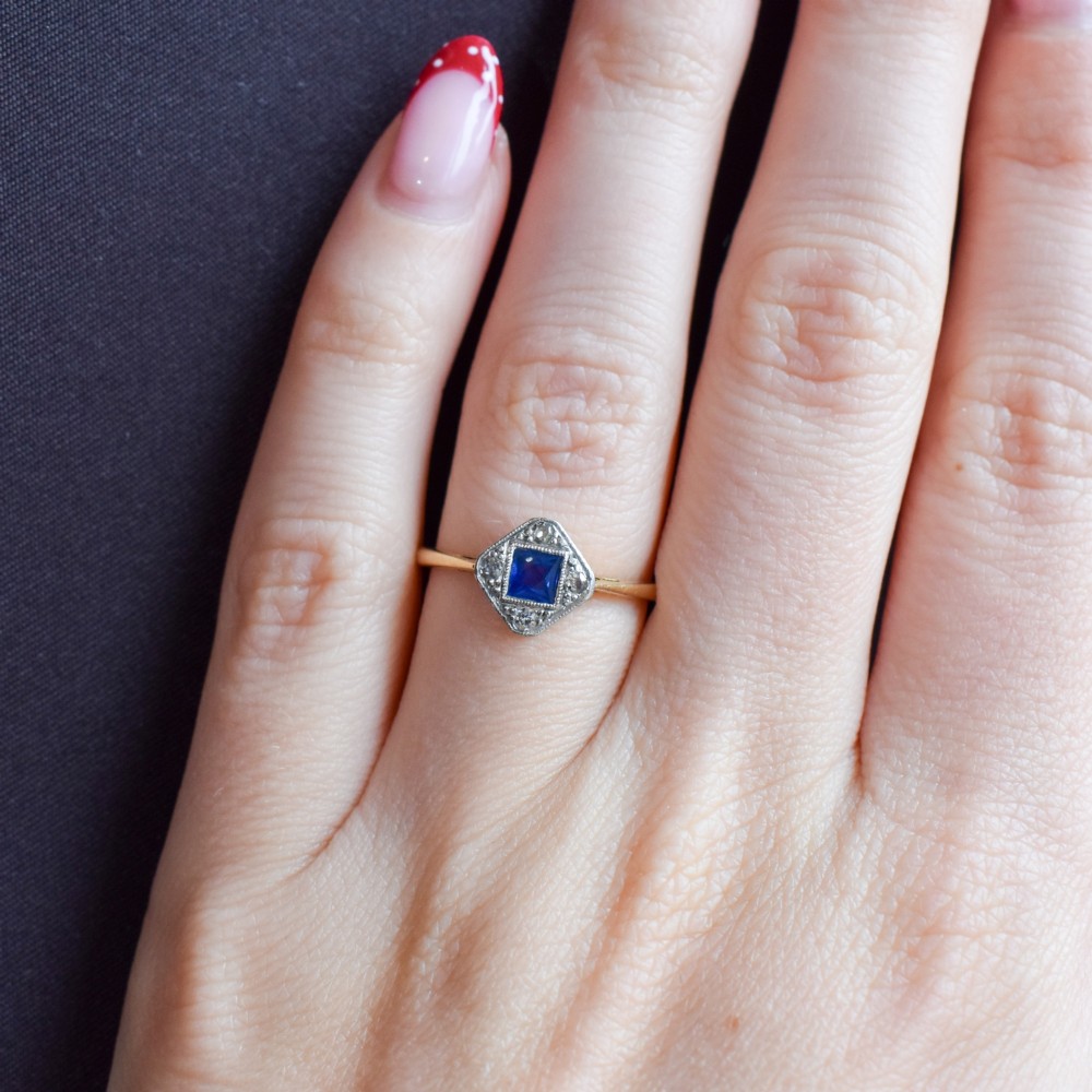 An Art Deco ring displaying a square blue sapphire surrounded by diamonds in a diamond-shaped geometric setting on an 18ct yellow gold band. View X