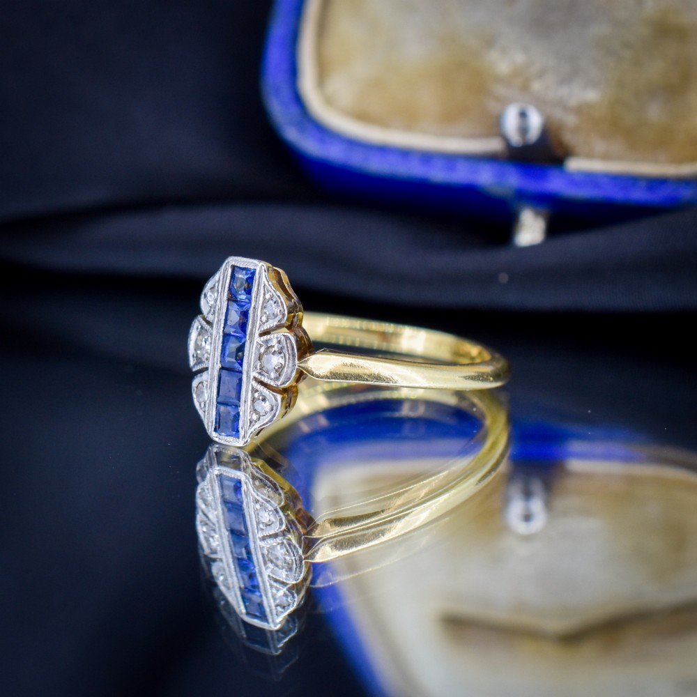 An Art Deco 18ct gold ring featuring a central vertical row of square blue sapphires flanked by diamond accents in a navette setting. View VI