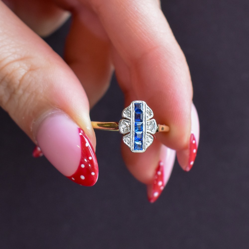 An Art Deco 18ct gold ring featuring a central vertical row of square blue sapphires flanked by diamond accents in a navette setting. View X