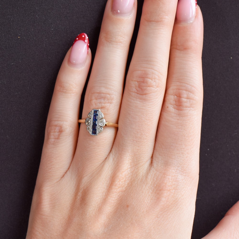 An Art Deco 18ct gold ring featuring a central vertical row of square blue sapphires flanked by diamond accents in a navette setting. View XI