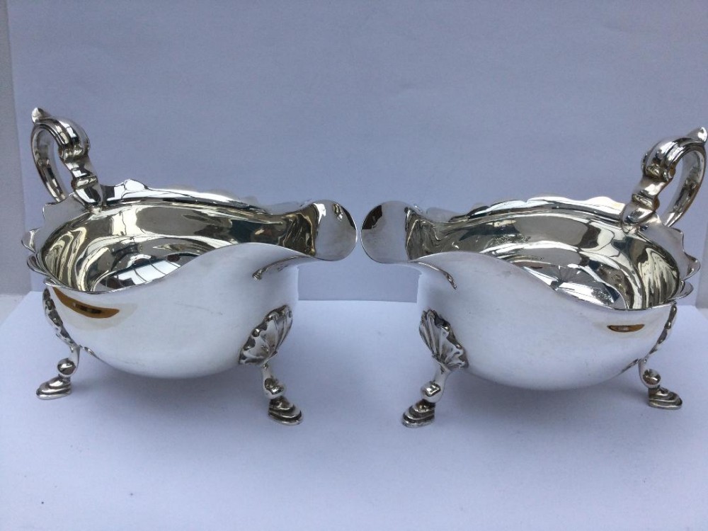 A matching pair of polished silver sauce boats featuring scalloped rims, scroll handles, and three shell-capped feet.