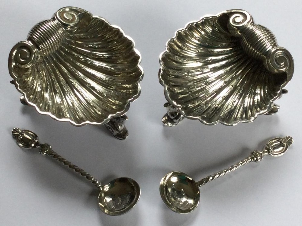 A pair of Victorian sterling silver shell-shaped salt cellars resting on cast feet alongside two matching silver spoons.